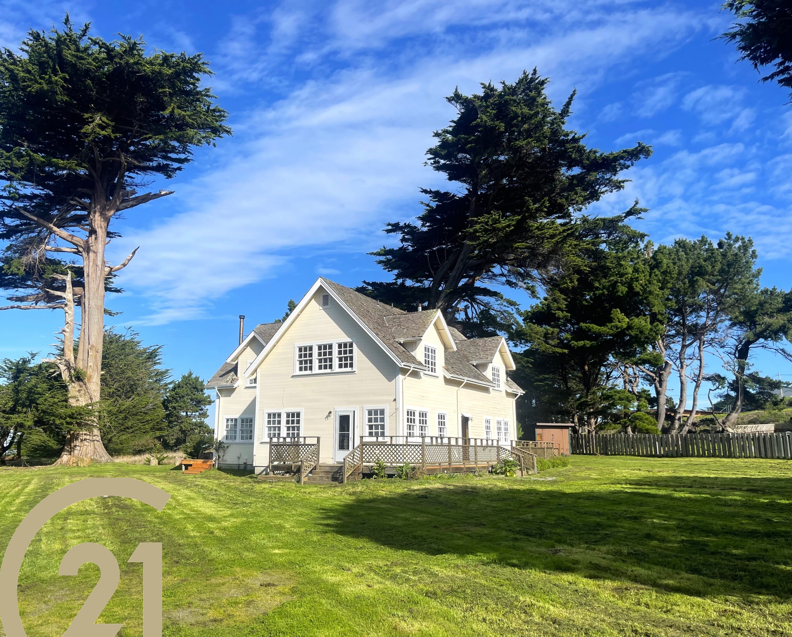 Two story gabled house with green lawn, blue sky and tall cypress trees in the background